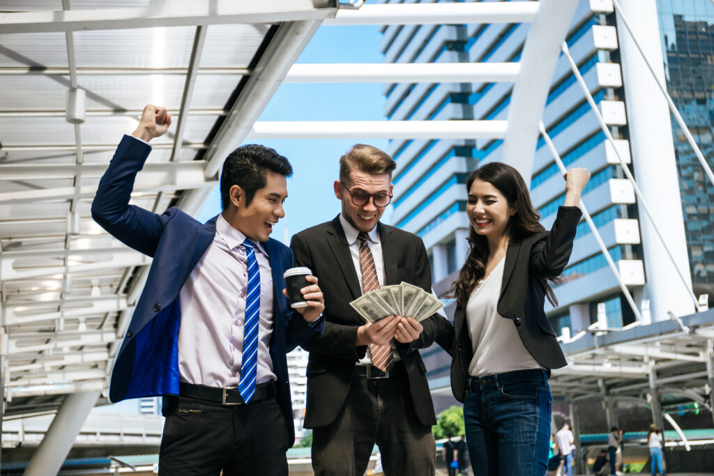 smiling business colleagues with arms raised holding currency standing outdoors